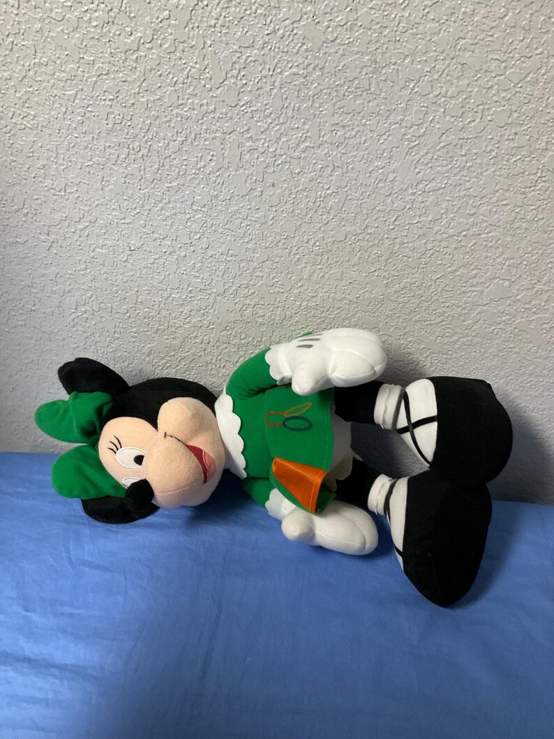 Puede incluir: Mu&ntilde;eca de peluche de Minnie Mouse con un atuendo verde con cuello blanco y lazo verde a juego. La mu&ntilde;eca tiene orejas negras, boca roja y guantes y zapatos blancos. La mu&ntilde;eca est&aacute; tumbada sobre una superficie azul.