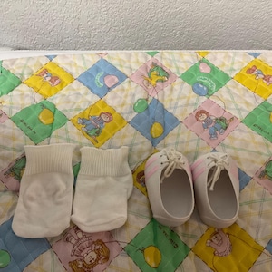 Może przedstawiać: Zestaw akcesoriów dla niemowląt, w tym dwie pary białych skarpet i para białych tenisówek z różowymi paskami. Przedmioty ułożone są na kolorowej kołdrze z powtarzającym się wzorem dzieci i zabawek.