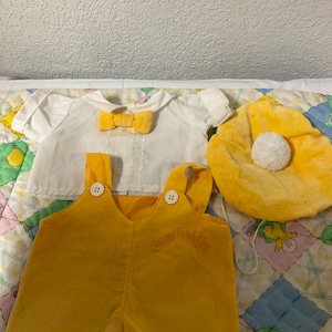 May include: A yellow and white outfit for a baby doll. The outfit includes a white shirt with a yellow bow tie, yellow dungarees with the text "Care Bears" embroidered on the front, and a yellow hat with a white pom-pom.