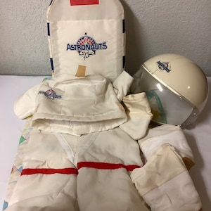 May include: A white astronaut costume for a child, including a helmet, jacket, pants, gloves, and a back pack. The costume has red accents and the text "Young Astronauts" on the back pack and helmet.