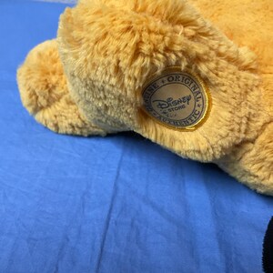 May include: A plush, golden-yellow stuffed animal with a Disney Store Authentic tag. The tag is a circular patch with the Disney Store logo and the words "Genuine Original Authentic". The toy is on a blue surface.