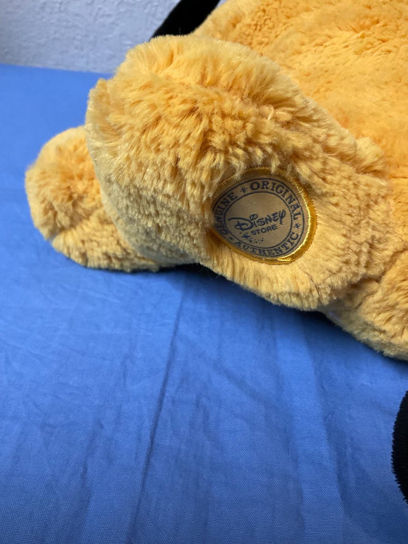 May include: A plush, golden-yellow stuffed animal with a Disney Store Authentic tag. The tag is a circular patch with the Disney Store logo and the words "Genuine Original Authentic". The toy is on a blue surface.