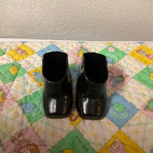 May include: A pair of black doll shoes with a shiny finish. The shoes are on a colourful quilted fabric.