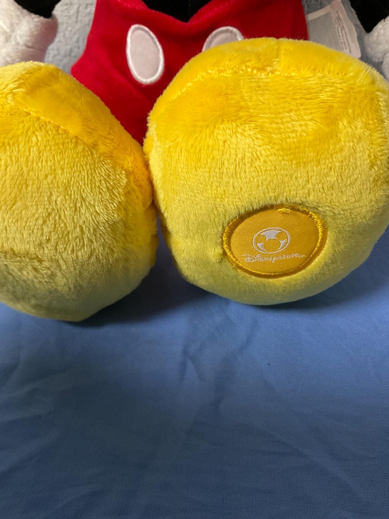 Pode incluir: Um brinquedo de pel&uacute;cia amarelo Mickey Mouse com uma camisa vermelha e contornos pretos. O brinquedo tem uma etiqueta amarela no p&eacute; direito com o logotipo da Disney.