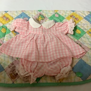 Puede incluir: Un conjunto de vestido y bombachos de vichy rosa y blanco para una muñeca. El vestido tiene un cuello blanco y encaje en las mangas. Los bombachos tienen encaje y lunares blancos.