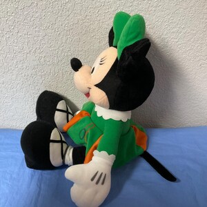 Puede incluir: Mu&ntilde;eca de peluche de Minnie Mouse sentada, vestida con un vestido verde con detalles en naranja y un lazo verde a juego. La mu&ntilde;eca tiene orejas negras, una cola negra y guantes blancos con tres l&iacute;neas negras en cada mano.