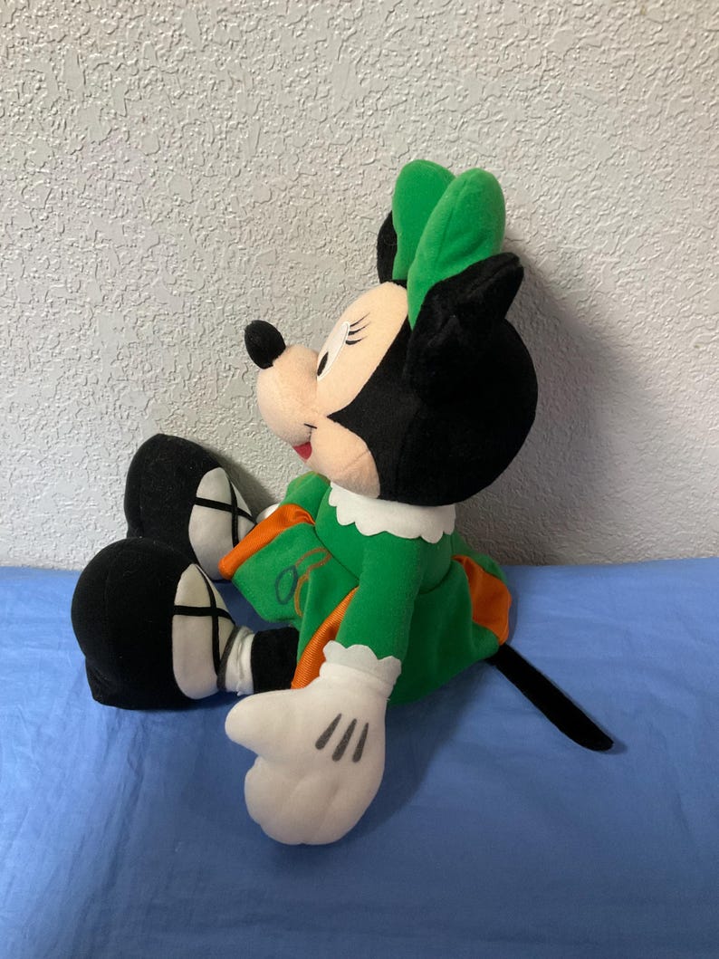 Puede incluir: Mu&ntilde;eca de peluche de Minnie Mouse sentada, vestida con un vestido verde con detalles en naranja y un lazo verde a juego. La mu&ntilde;eca tiene orejas negras, una cola negra y guantes blancos con tres l&iacute;neas negras en cada mano.