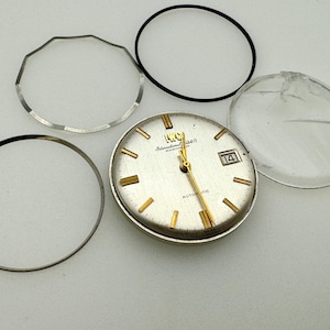 Può includere: Un quadrante argentato con lancette e indicatori dorati, che mostra la data. L'orologio è circondato da varie parti dell'orologio, tra cui un cristallo trasparente e incrinato e diversi anelli neri e argentati. Il quadrante recita "IWC International Watch Co. Automatic."