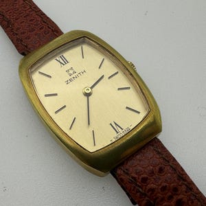 Pode incluir: Um relógio Zenith vintage com caixa retangular dourada e mostrador dourado claro. O relógio apresenta marcadores de hora e ponteiros pretos, com o logotipo Zenith e o texto "SWISS MADE". Possui uma pulseira de couro marrom.