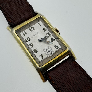 Pode incluir: Relógio de pulso retangular vintage com caixa dourada e pulseira de couro castanho. O mostrador do relógio é branco com algarismos e ponteiros pretos. O mostrador do relógio tem o nome da marca "Silvana" impresso.