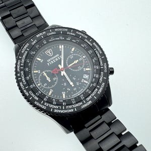 Pode incluir: Relógio DETOMASO FIRENZE preto com pulseira de metal preta. O mostrador é preto com detalhes brancos e vermelhos, incluindo um taquímetro e mostradores secundários. A luneta exibe nomes de cidades.
