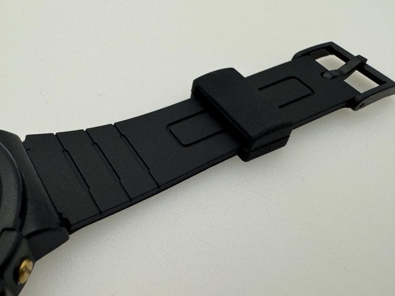 May include: Black watch strap with a buckle closure. The strap is made of a textured rubber material.