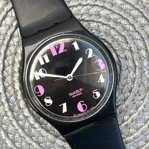 Può includere: Un orologio Swatch nero con cinturino nero e quadrante nero. I numeri sul quadrante sono rosa e bianchi. L'orologio dice "Swatch Swiss" sul quadrante.