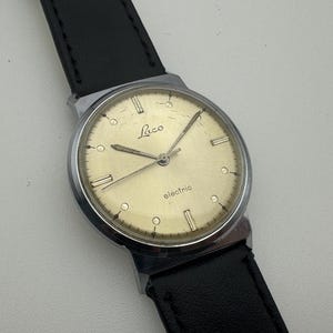 May include: A vintage Laco electric wristwatch with a silver-tone case and a cream-colored dial. The dial features silver hands and markers, with the word "electric" printed below the center. The watch has a black leather strap.