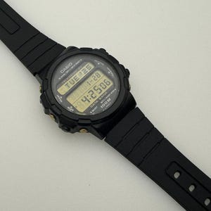 May include: Black Casio digital wristwatch with a black band and gold accents. The watch face displays the time, date, and day of the week. The watch is water resistant to 100 meters.