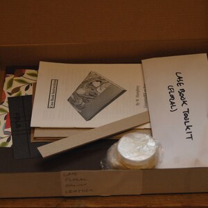 May include: A brown cardboard box with a white lid is open, revealing a case book toolkit. The toolkit includes instructions, a patterned fabric, a piece of cardboard, a ruler, a small jar of glue, and a white envelope with the text "Case Book Toolkit (Floral)".