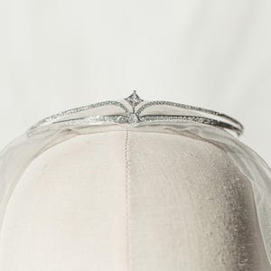 Pode incluir: Uma tiara de tom prateado com um design de banda dupla incrustada com pequenos cristais transparentes. O centro da tiara é acentuado com um cristal único e proeminente de corte quadrado. A peça é colocada sobre um véu de tecido branco.