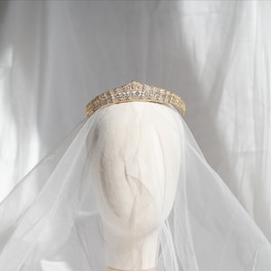 May include: A gold-toned tiara featuring a series of clear, sparkling stones arranged in a pointed, symmetrical pattern. The tiara is positioned atop a plain, neutral-colored head form, with a sheer, white, translucent veil draped over it.
