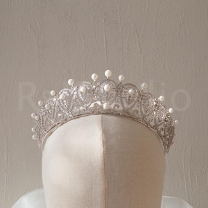 May include: A silver tiara with a delicate design and pearl accents. The tiara is on a white mannequin head.