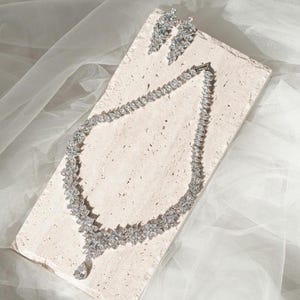 Puede incluir: Un conjunto de collar y pendientes plateados. El collar presenta un diseño floral con un colgante en forma de lágrima. Los pendientes tienen un diseño floral similar. Las joyas se exhiben sobre una superficie clara y texturizada.