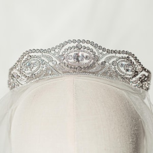 May include: A silver tiara with a delicate, ornate design, featuring numerous sparkling clear gemstones. The tiara is set on a white form, with a sheer veil draped below. The design includes oval and circular patterns.