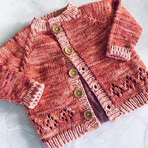 Puede incluir: Cárdigan tejido a mano en tonos rojos, naranjas y rosas. El suéter con botones presenta un patrón de punto texturizado, botones de madera y calados decorativos en el borde inferior. Las mangas y el cuello están acabados con un ribete de canalé.