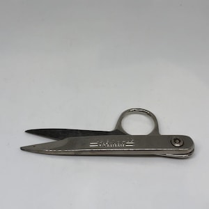 Könnte beinhalten: Ein silberfarbener Fadenabschneider aus Metall mit einem einzelnen runden Fingerring und spitzen Klingen. Auf der Seite des Werkzeugs befindet sich eine Gravur mit dem Schriftzug "CLOVER" sowie weitere kleine, teilweise sichtbare Buchstaben.