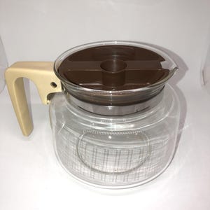 May include: A clear glass coffee carafe with a brown lid and a cream-colored handle. The carafe has a stainless steel band around the middle and a spout for pouring. The design is simple and functional, suitable for brewing coffee.