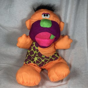 May include: A cartoon caveman plush toy. The toy is predominantly orange, with a purple mouth, pink tongue, and green nose. It features a green and brown patterned loincloth and black hair.
