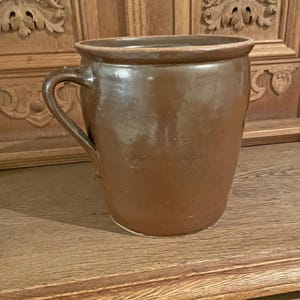 May include: A large, brown earthenware crock with a single handle. The crock has a wide mouth and a rounded body.