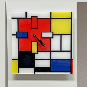 May include: A clock with a black face and hands, featuring a geometric design with red, yellow, blue, and white squares. The clock is made of glass and has a modern, abstract look.