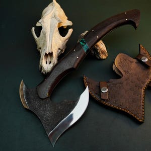 May include: A handcrafted axe with a dark wooden handle, accented with brass pins and a green stone inlay. The axe head is made of metal with a polished blade. A brown leather sheath with embossed design is included.