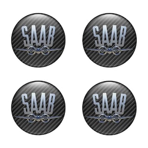 Könnte beinhalten: Vier runde schwarze und silberne Autoembleme mit einem Karbonfaser-Muster. Jedes Emblem zeigt das Wort "SAAB" in silbernen Buchstaben und ein stilisiertes Flugzeugpropeller-Design.