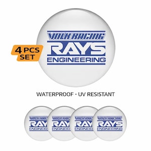 May include: A set of four white, round stickers with blue text. The stickers feature the text "VOLK RACING RAYS ENGINEERING". The set includes a yellow and orange label that says "4 PCS SET". The stickers are waterproof and UV resistant.