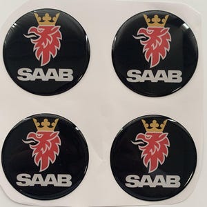Könnte beinhalten: Vier schwarze, kreisförmige Aufkleber mit dem Wort "SAAB" in Weiß. Jeder Aufkleber zeigt ein rotes Greifenemblem mit einer goldenen Krone. Die Aufkleber befinden sich auf einem weißen Hintergrund.