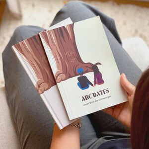 Könnte beinhalten: Zwei Bücher mit weißen Einbänden werden gehalten. Das obere Buch trägt den Titel "ABC DATES" mit einer Illustration von zwei Figuren unter einem Baum. Das andere Buch hat eine braune Baumillustration und die Worte "You & Me".