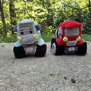 May include: Two handmade crochet toy trucks. One is light blue and gray, the other is red and gray. Both have black wheels and small tags. The toys are sitting on a concrete surface with a blurred green background.