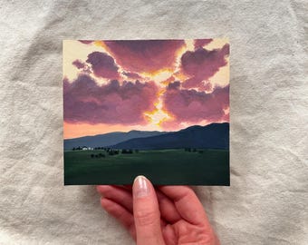 May include: A small square painting depicts a sunset over a landscape. The sky is filled with pink and purple clouds, with a bright yellow light breaking through. Below, dark green mountains and a field are visible.