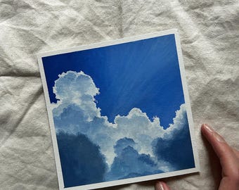 May include: Square painting of a blue sky with fluffy white and gray clouds. The artwork features a gradient of blues, with lighter shades at the top and darker shades at the bottom. The painting is held by a person.