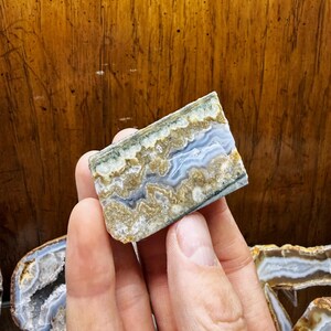 May include: A hand holds a rectangular, polished agate stone. The stone features layers of blue, tan, and white, with a wavy pattern. The edges show a mix of green and brown. Other agate slices are visible in the background.