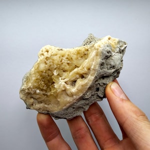 Może przedstawiać: Naturalny, surowy okaz minerału z jasnożółtym, krystalicznym wnętrzem, zamkniętym w szarej, teksturowanej matrycy. Minerał ma szorstką, nierówną powierzchnię z widocznymi formacjami i zmienną kolorystyką i fakturą. Okaz jest trzymany przez dłoń.