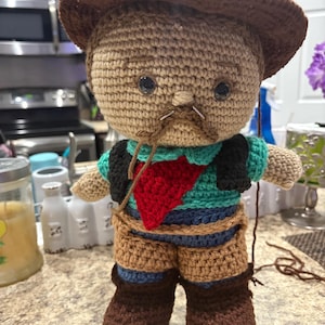 May include: A handmade crochet doll dressed as a cowboy, featuring a brown hat, vest, and boots. The doll has a red neckerchief and a mustache. The doll is made with various shades of brown, blue, green, and black yarn.