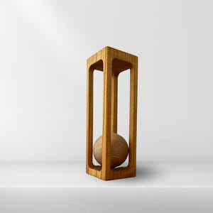 Op de afbeelding: Een houten sculptuur met een bol in een rechthoekig frame. De sculptuur is gemaakt van lichtbruin hout en heeft een natuurlijke afwerking.