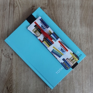 Puede incluir: Una libreta azul con un marcapáginas de tela blanca con un patrón de libros coloridos y un bolígrafo rojo. El marcapáginas tiene el texto "TOTAL THIS BOOK".