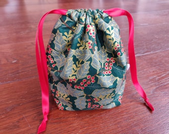 Pode incluir: Uma bolsa de cordão verde com um padrão de azevinho e bagas. A bolsa tem laços de fita vermelha.