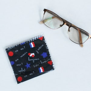 Puede incluir: Gafas con montura de metal marrón y lentes transparentes sobre una superficie blanca. Un paño de limpieza negro con frases francesas y gráficos de banderas está en un envoltorio de plástico transparente.