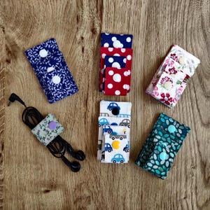 May include: Six rectangular fabric cord organizers with snap closures. Patterns include blue leaves, red and blue polka dots, floral prints, blue cars on white, and green ovals. One organizer is wrapped around black wired earbuds with a purple snap.