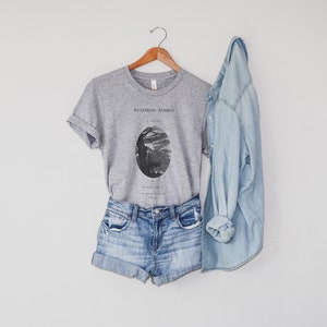 May include: A gray t-shirt with a black and white illustration of a tree and the text "Wuthering Heights" on the front. The t-shirt is paired with denim shorts and a light blue denim jacket.