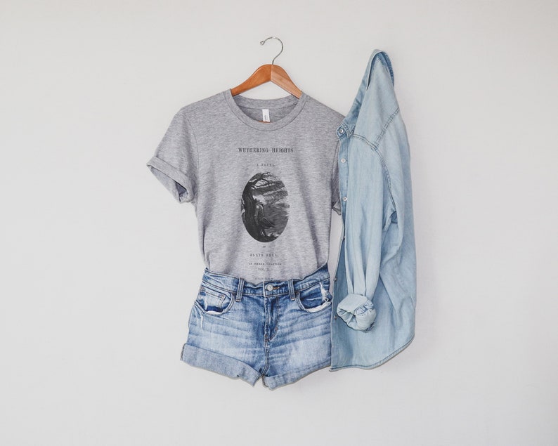 May include: A gray t-shirt with a black and white illustration of a tree and the text "Wuthering Heights" on the front. The t-shirt is paired with denim shorts and a light blue denim jacket.