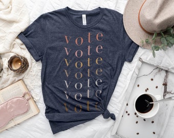 May include: A navy blue t-shirt with the word "vote" repeated in a stacked pattern in different shades of pink, orange, and gold.